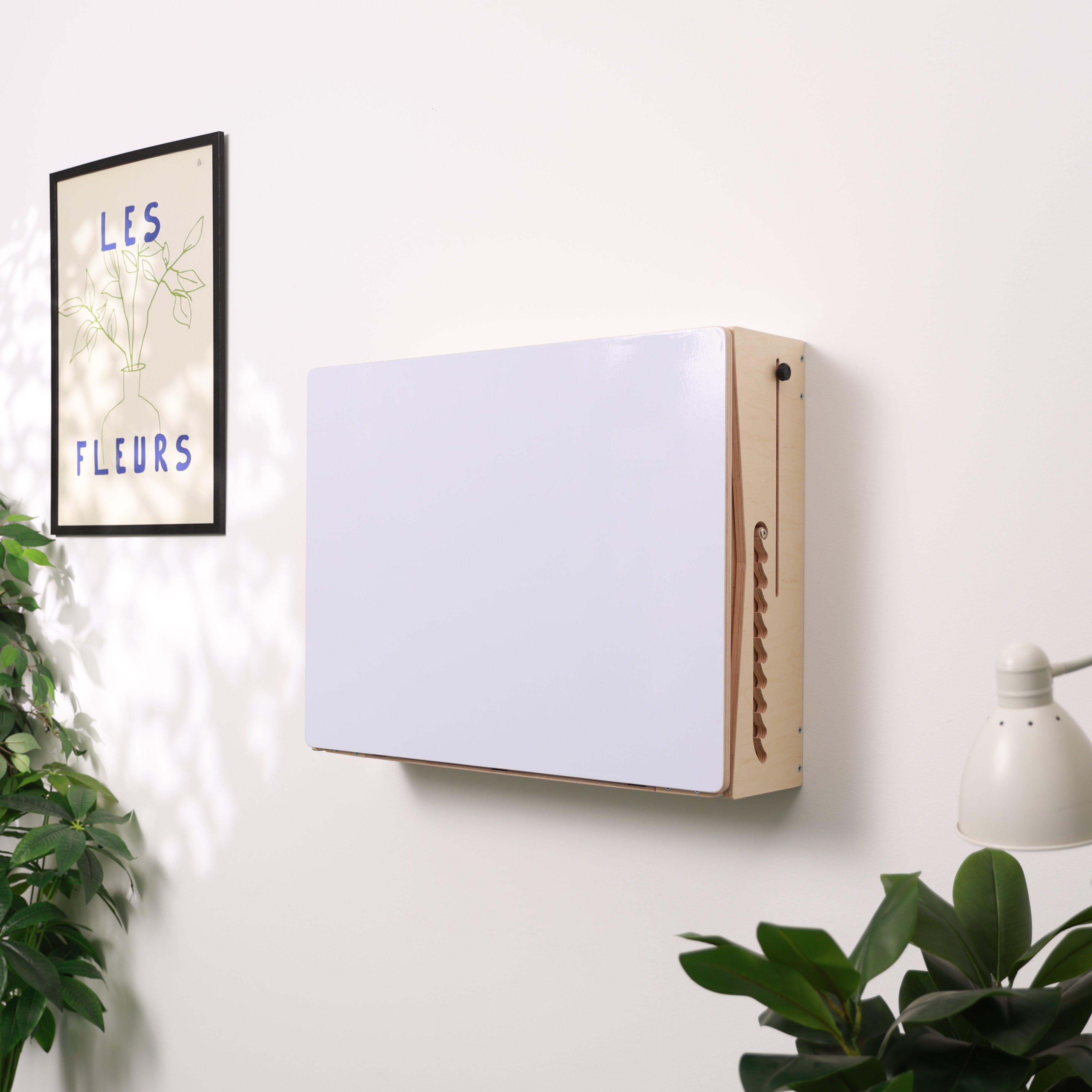 A wall-mounted rectangular whiteboard with a light wood frame, shown on a white wall in a styled room with plants, a framed print, and a lamp.