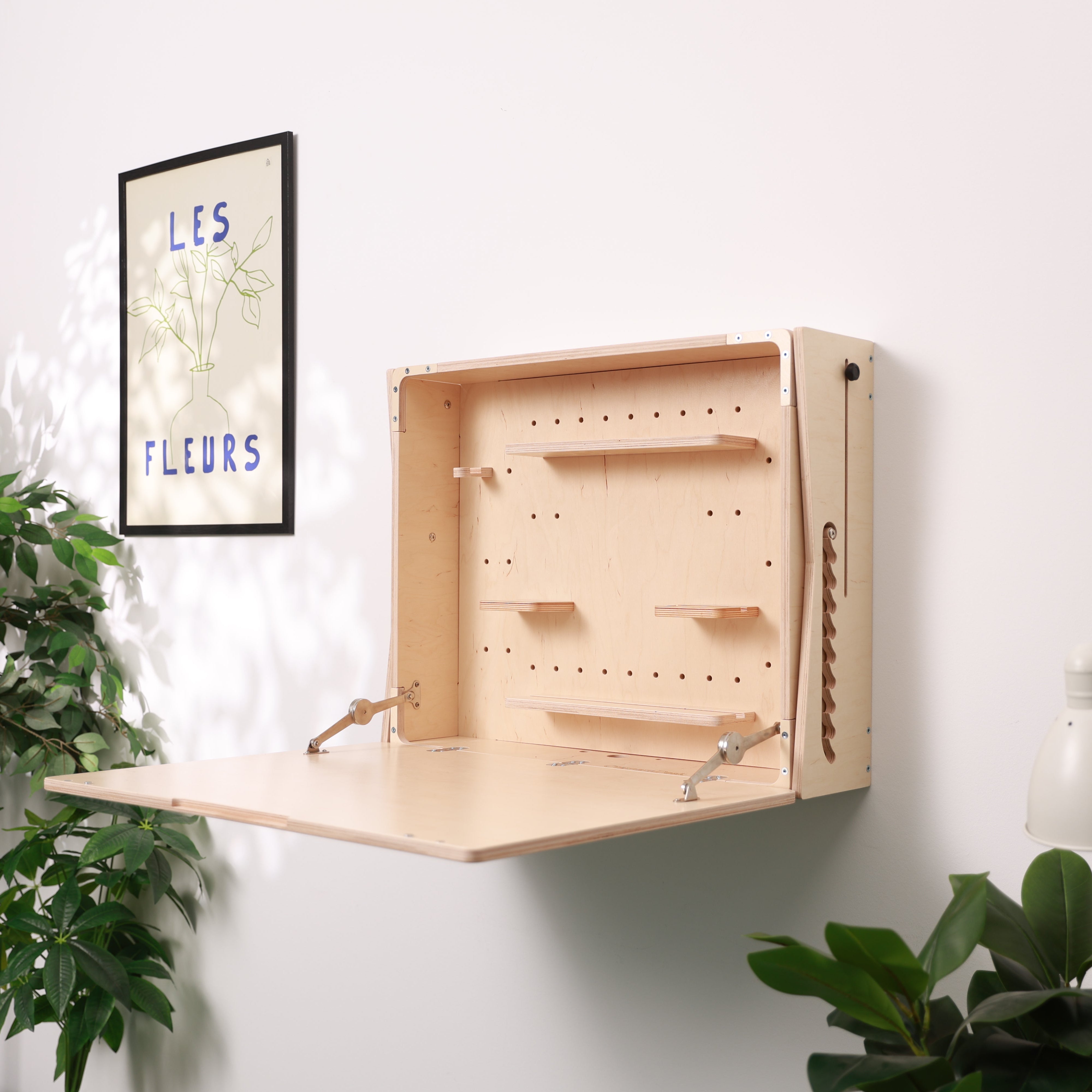 A light natural wood wall-mounted desk or fold-out table unit with an open front and pegboard-style interior, shown mounted on a white wall in a styled room with framed wall art, plants, and a lamp.