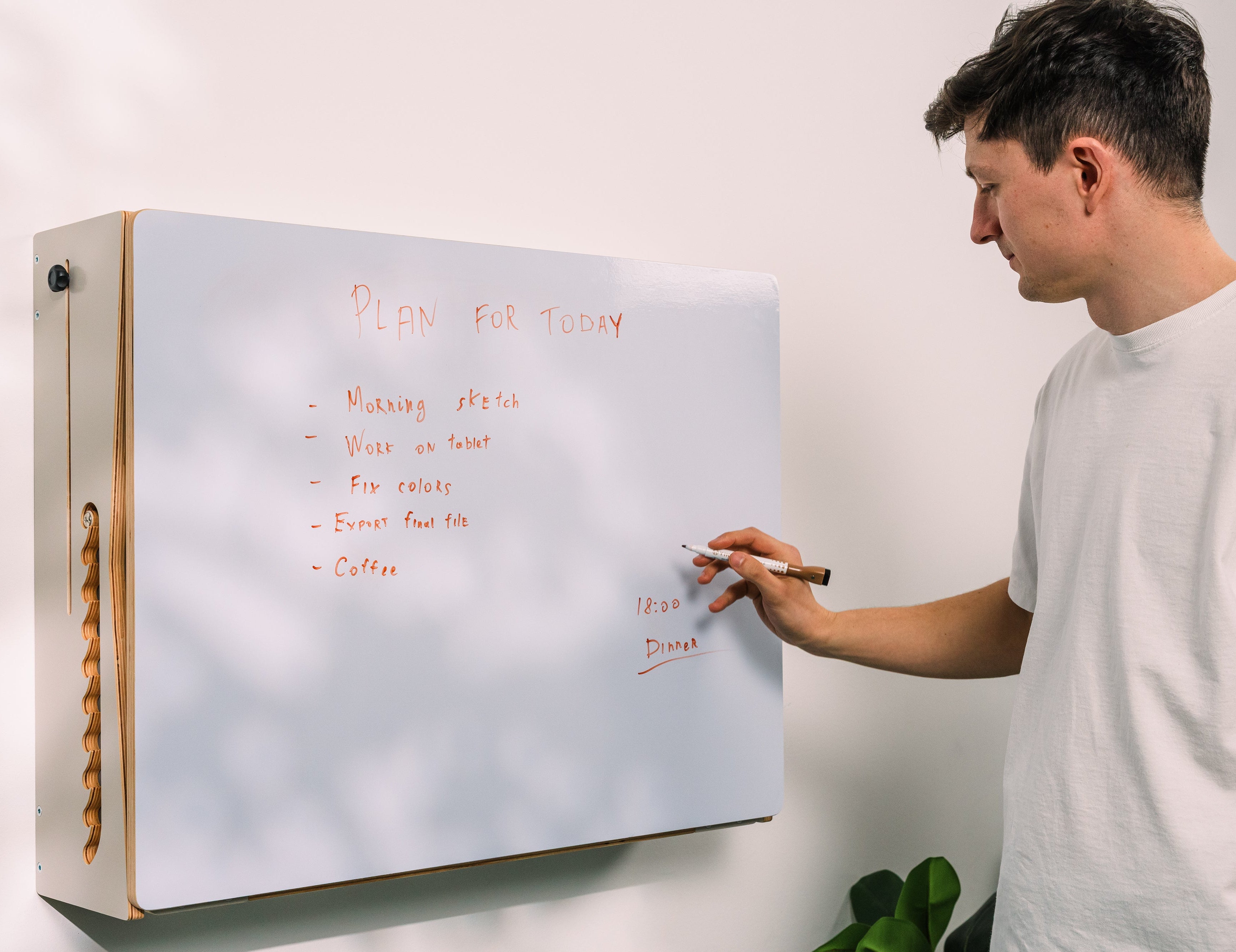 A man in a white t-shirt stands next to a white magnetic dry-erase board, writing on it with a marker. The board has a light wood side frame and is mounted on a white wall.